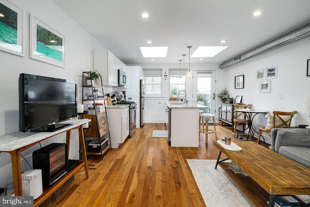 kitchen with light wood-type flooring, a skylight, stainless steel appliances, a kitchen island with sink, and hanging light fixtures