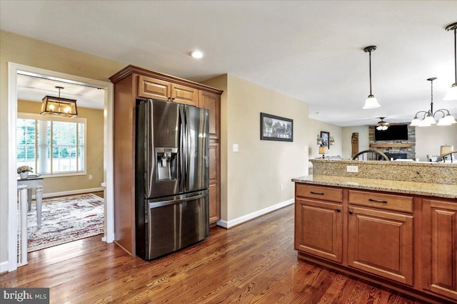 kitchen with light stone counters, ceiling fan with notable chandelier, a brick fireplace, stainless steel fridge with ice dispenser, and dark hardwood / wood-style flooring