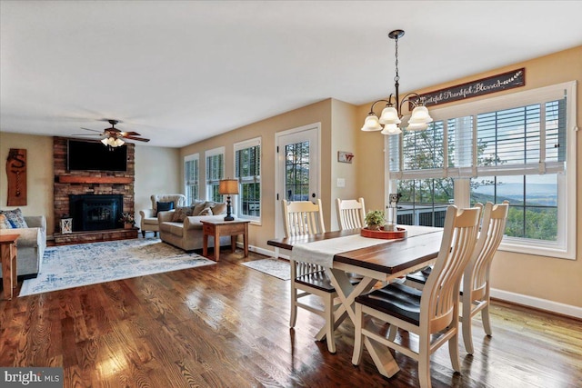 dining room featuring a stone fireplace, hardwood / wood-style floors, a healthy amount of sunlight, and ceiling fan with notable chandelier