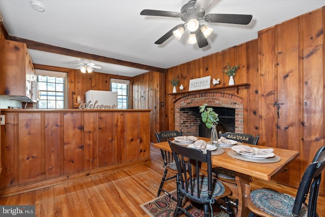 dining room with ceiling fan, light wood-type flooring, a brick fireplace, beamed ceiling, and wooden walls