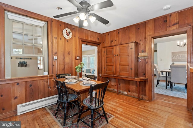 dining space with light hardwood / wood-style flooring, a baseboard radiator, wooden walls, and ceiling fan with notable chandelier