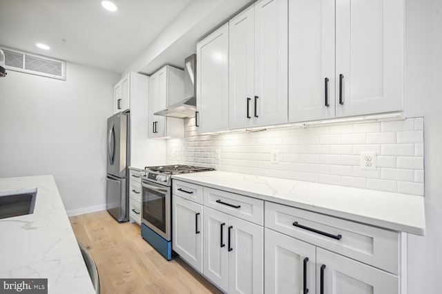 kitchen with wall chimney range hood, stainless steel appliances, light stone countertops, white cabinets, and light hardwood / wood-style floors