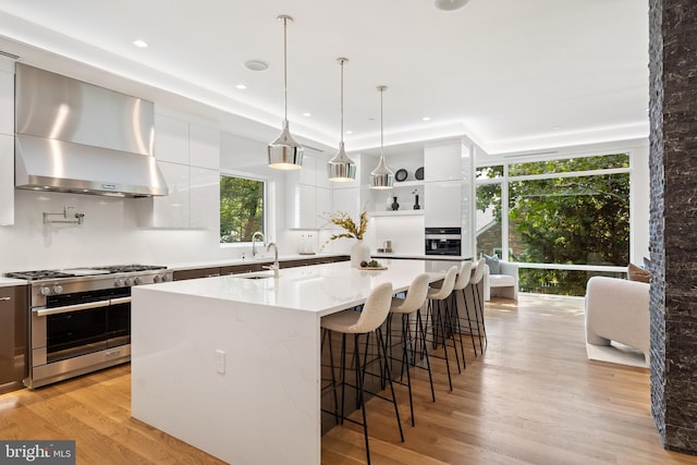 kitchen with wall chimney exhaust hood, a kitchen island with sink, light wood-type flooring, stainless steel stove, and white cabinetry