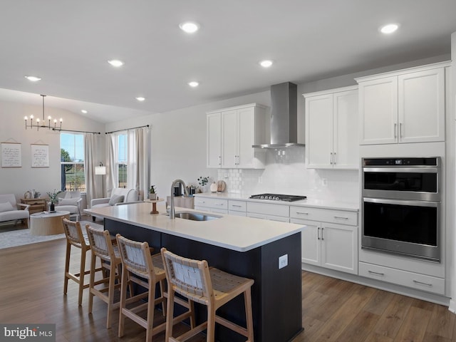 kitchen with stainless steel appliances, wall chimney range hood, dark hardwood / wood-style flooring, and an island with sink