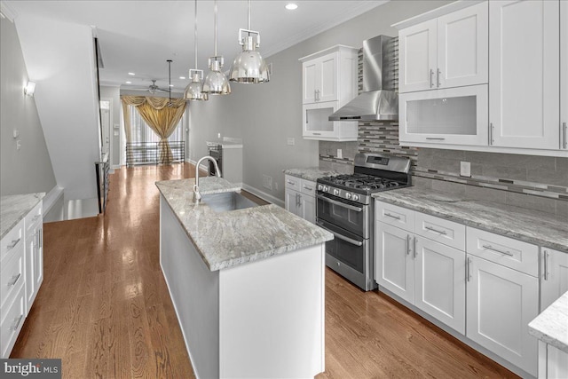 kitchen featuring wall chimney exhaust hood, gas stove, a center island with sink, sink, and white cabinetry