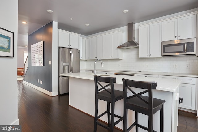 kitchen featuring wall chimney exhaust hood, dark hardwood / wood-style flooring, an island with sink, appliances with stainless steel finishes, and white cabinetry