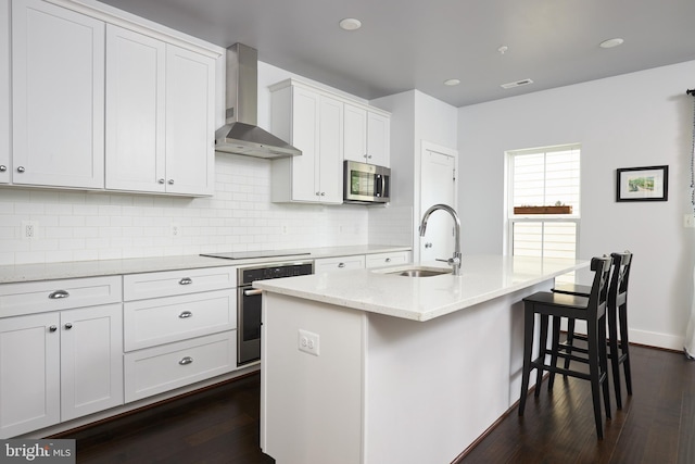 kitchen with stainless steel appliances, dark wood-type flooring, sink, wall chimney range hood, and a center island with sink