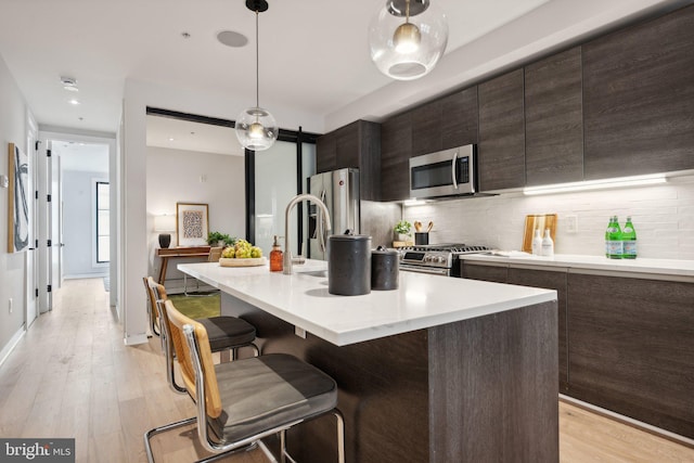 kitchen with a kitchen island with sink, stainless steel appliances, backsplash, pendant lighting, and light hardwood / wood-style floors