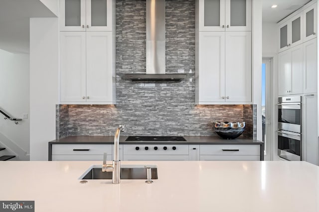 kitchen with decorative backsplash, stainless steel double oven, sink, wall chimney range hood, and white cabinets