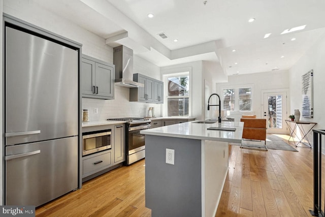 kitchen with appliances with stainless steel finishes, wall chimney range hood, gray cabinets, a kitchen island with sink, and light hardwood / wood-style flooring