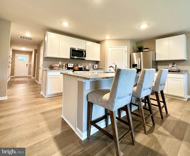 kitchen with light stone counters, stainless steel appliances, white cabinets, light hardwood / wood-style flooring, and a center island with sink