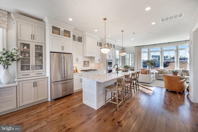 kitchen with white cabinets, dark wood-type flooring, appliances with stainless steel finishes, decorative light fixtures, and a center island with sink