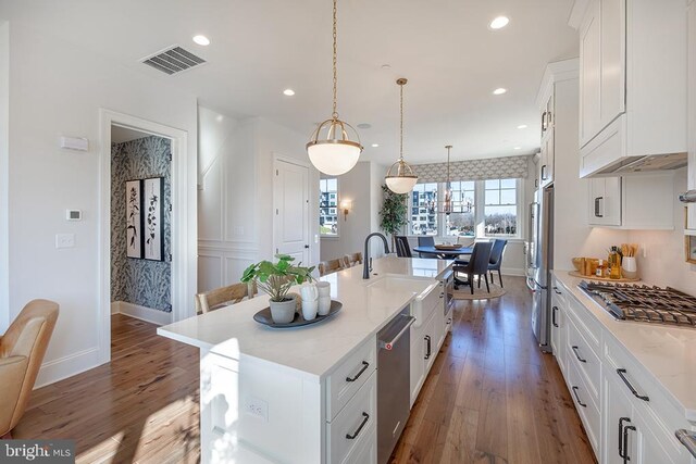 kitchen with dark hardwood / wood-style flooring, stainless steel appliances, hanging light fixtures, white cabinets, and an island with sink