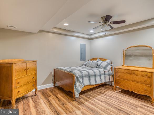 bedroom with ceiling fan, a raised ceiling, wood-type flooring, and electric panel