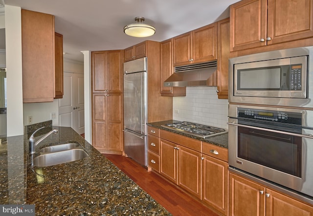 kitchen with sink, backsplash, dark hardwood / wood-style flooring, dark stone counters, and built in appliances