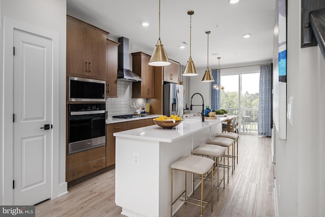 kitchen featuring wall chimney range hood, appliances with stainless steel finishes, a kitchen bar, pendant lighting, and a kitchen island with sink