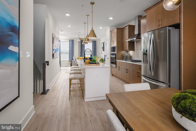 kitchen with wall chimney exhaust hood, a center island with sink, pendant lighting, appliances with stainless steel finishes, and light hardwood / wood-style floors