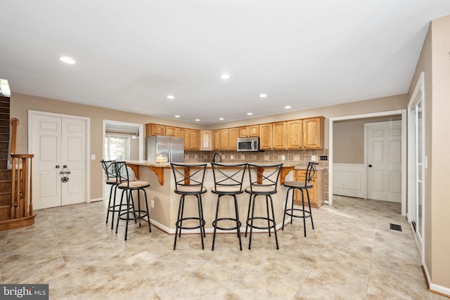 kitchen with a center island with sink, backsplash, light stone countertops, a kitchen bar, and stainless steel appliances