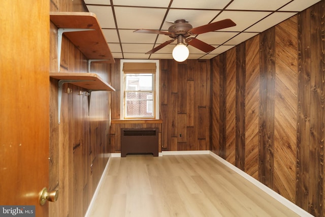 interior space with a drop ceiling, radiator heating unit, wooden walls, ceiling fan, and wood-type flooring