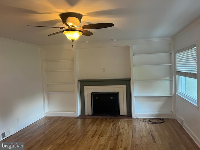 unfurnished living room with built in shelves, crown molding, wood-type flooring, and ceiling fan