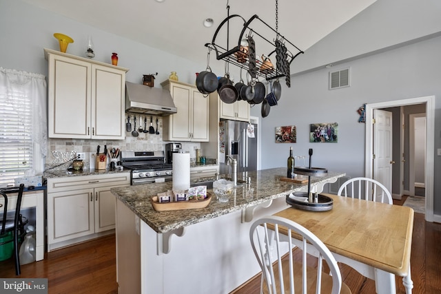 kitchen featuring wall chimney range hood, a kitchen island with sink, dark stone counters, lofted ceiling, and appliances with stainless steel finishes