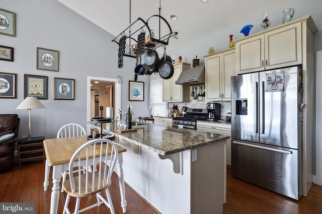 kitchen with cream cabinets, wall chimney exhaust hood, dark wood-type flooring, appliances with stainless steel finishes, and dark stone countertops