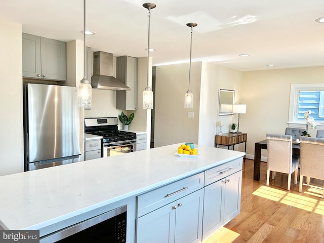 kitchen with wall chimney exhaust hood, hanging light fixtures, appliances with stainless steel finishes, light stone counters, and light hardwood / wood-style floors