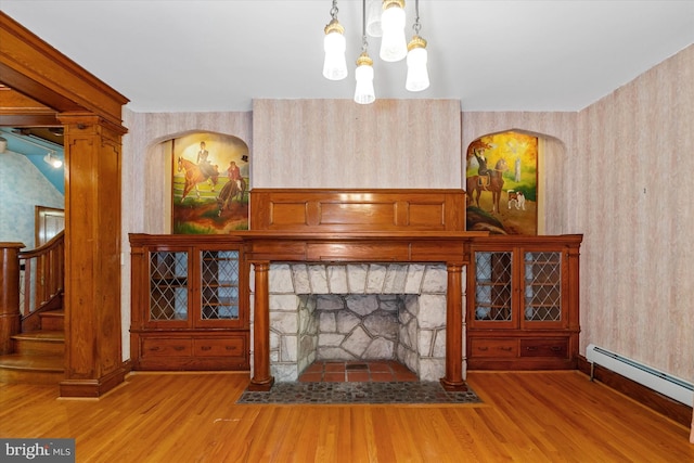 living room featuring a stone fireplace, baseboard heating, wallpapered walls, wood finished floors, and stairs
