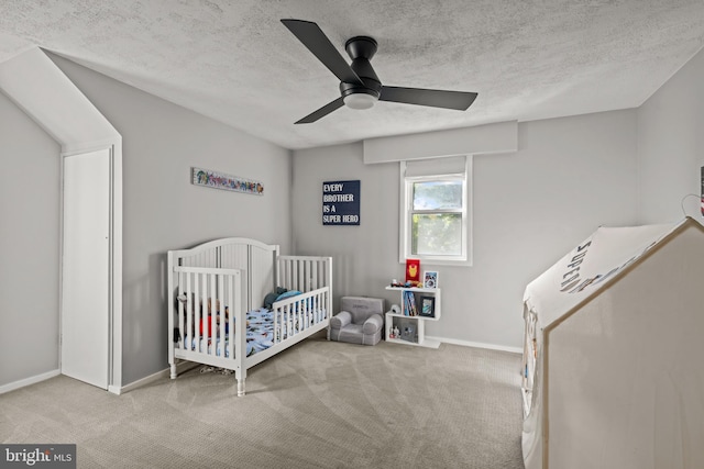 carpeted bedroom featuring a textured ceiling, a nursery area, baseboards, and a ceiling fan