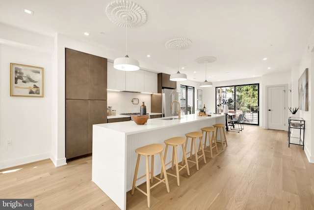 kitchen with an island with sink, hanging light fixtures, high quality fridge, white cabinets, and light hardwood / wood-style floors