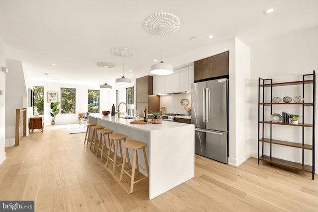 kitchen with stainless steel fridge, hanging light fixtures, dark brown cabinets, an island with sink, and light hardwood / wood-style flooring