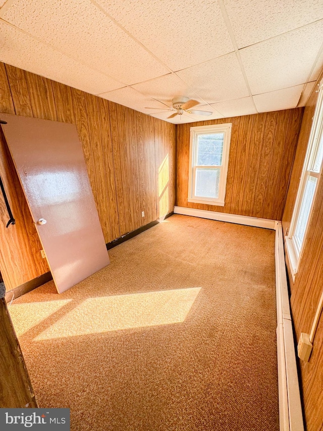 carpeted empty room with a drop ceiling, wood walls, and ceiling fan