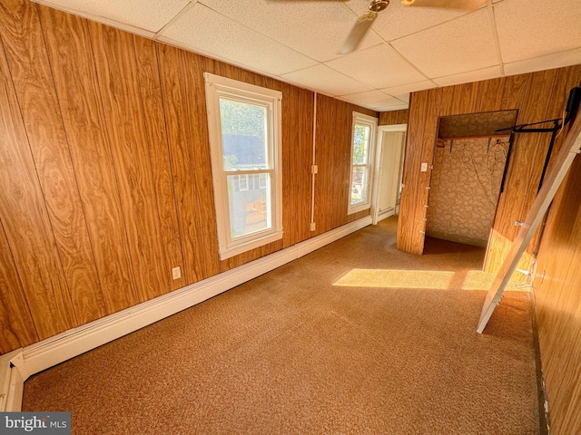 carpeted spare room featuring a paneled ceiling, wooden walls, a baseboard radiator, and ceiling fan