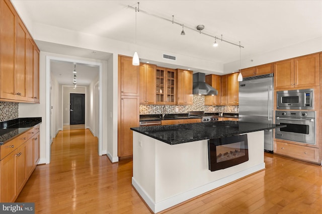 kitchen featuring a kitchen island, dark stone counters, hanging light fixtures, built in appliances, and wall chimney range hood