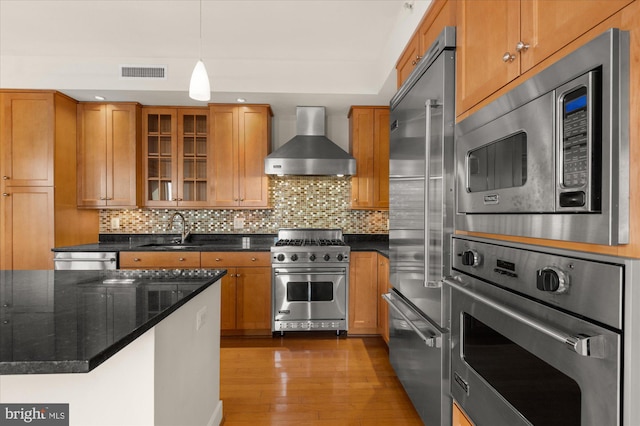 kitchen featuring sink, dark stone countertops, hanging light fixtures, built in appliances, and wall chimney range hood