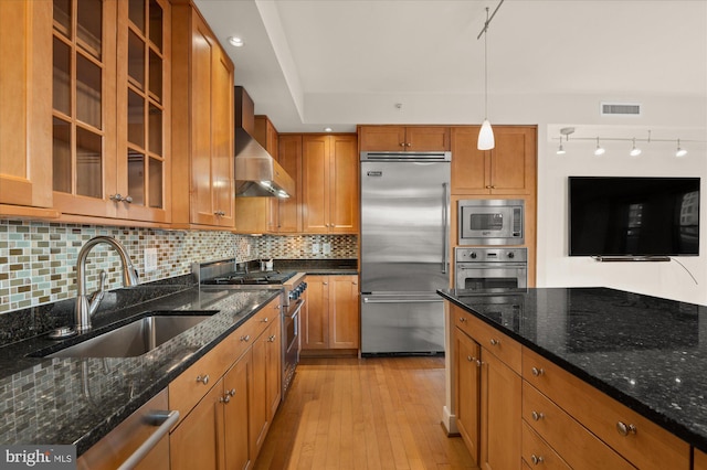 kitchen with decorative light fixtures, sink, dark stone countertops, built in appliances, and wall chimney exhaust hood