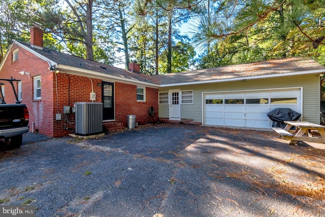 back of house featuring a chimney, aphalt driveway, central AC unit, brick siding, and a garage