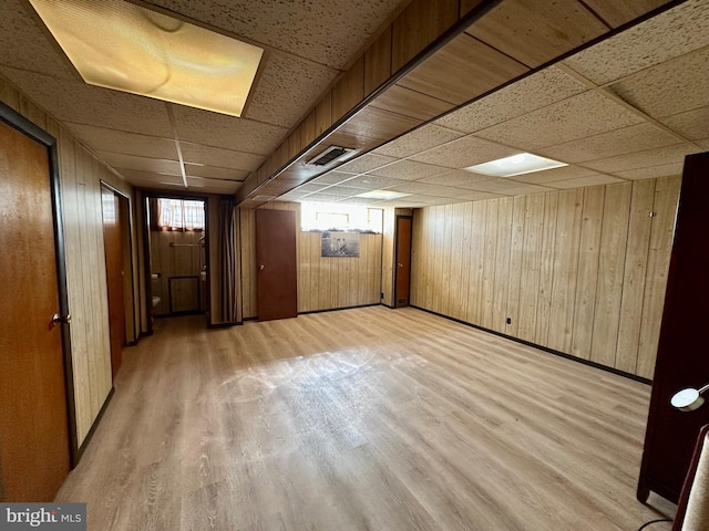 basement with a drop ceiling, wood walls, and light wood-type flooring