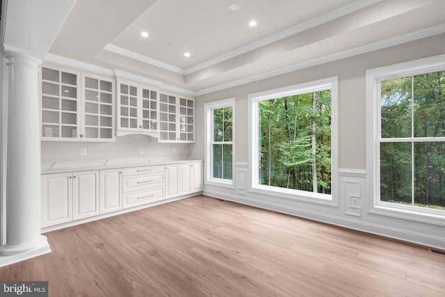 unfurnished dining area with ornamental molding, ornate columns, a tray ceiling, and light wood-type flooring