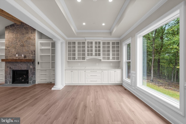 interior space with a stone fireplace, ornate columns, a tray ceiling, and light wood-type flooring