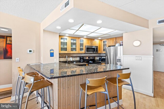 kitchen with appliances with stainless steel finishes, kitchen peninsula, light hardwood / wood-style floors, dark stone counters, and a breakfast bar area