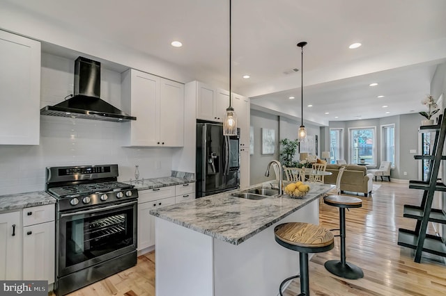 kitchen featuring white cabinetry, sink, wall chimney exhaust hood, stainless steel range with gas cooktop, and black refrigerator with ice dispenser