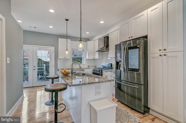 kitchen with stainless steel refrigerator with ice dispenser, wall chimney exhaust hood, a kitchen island with sink, black gas stove, and white cabinetry