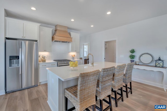 kitchen with an island with sink, white cabinets, custom range hood, appliances with stainless steel finishes, and a kitchen bar