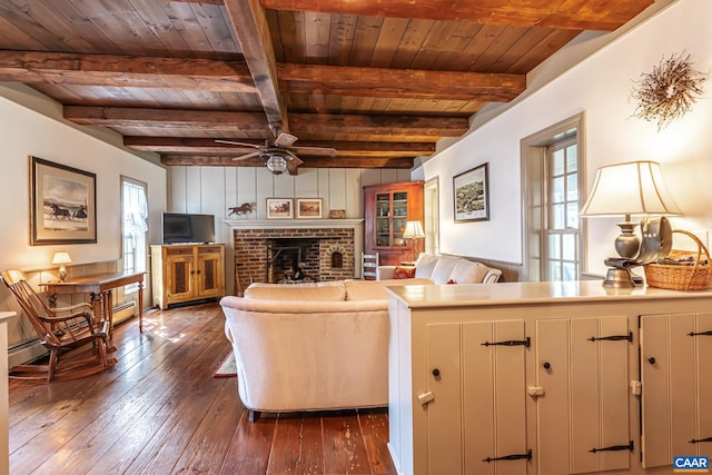 living room with a brick fireplace, beamed ceiling, ceiling fan, wood ceiling, and dark wood-type flooring