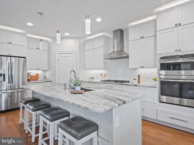 kitchen featuring wall chimney exhaust hood, appliances with stainless steel finishes, a kitchen island with sink, and hardwood / wood-style flooring