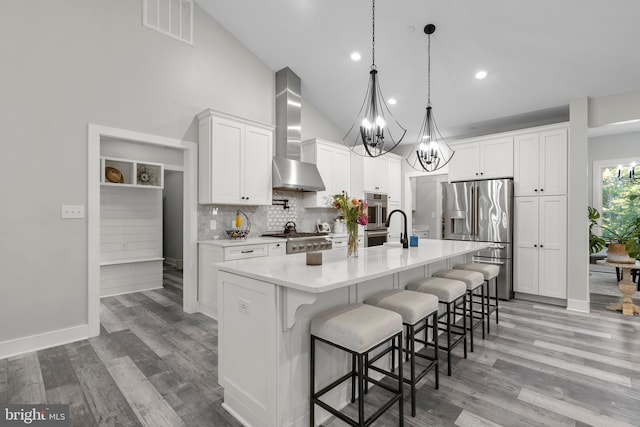 kitchen with a kitchen island with sink, wall chimney exhaust hood, white cabinets, appliances with stainless steel finishes, and light hardwood / wood-style floors