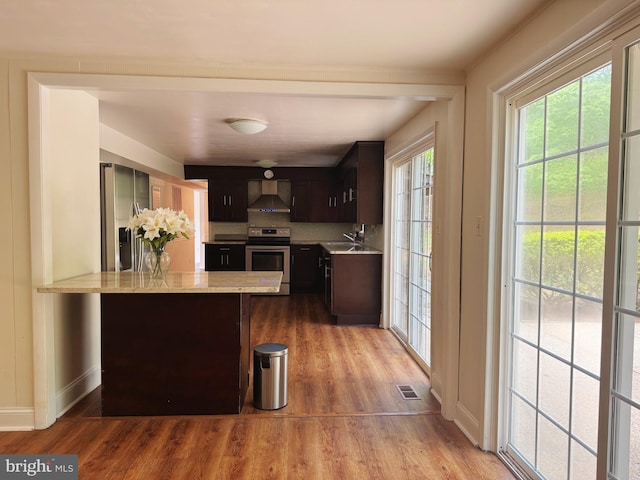 kitchen with dark brown cabinetry, wall chimney exhaust hood, stainless steel appliances, a peninsula, and light wood-type flooring