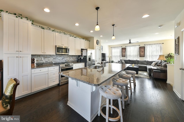 kitchen with appliances with stainless steel finishes, dark hardwood / wood-style flooring, dark stone counters, pendant lighting, and a kitchen island with sink