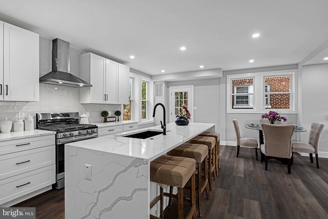 kitchen with wall chimney range hood, dark hardwood / wood-style floors, gas range, an island with sink, and sink